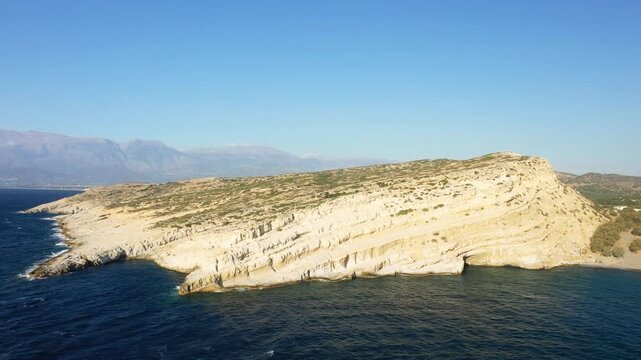 Aerial view of the iconic caves and rugged limestone cliffs along the coast of Matala, Crete, with clear blue sea and distant mountains under a clear sky.