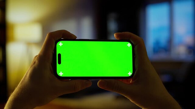 Hands holding smartphone with bright green chroma key screen in dimly lit cozy indoor room, ready for video replacement, app design preview or digital content editing