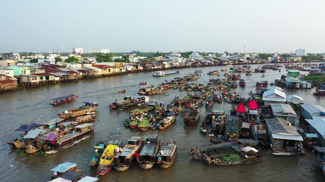 Aerial view of the bustling Cai Rang floating market with numerous boats trading goods on a wide river, surrounded by riverside buildings in Vietnam.
