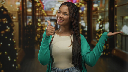 Fototapeta premium Woman points finger to palm in decorated building corridor wearing green cardigan and jeans, presenting open hand to camera with a smile; holiday joy.