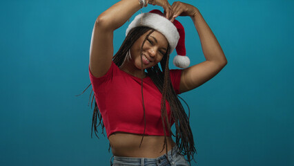 Fototapeta premium Woman forming a heart with her hands over head, showing midriff and wearing a santa hat in studio with teal backdrop; holiday cheer joyful.