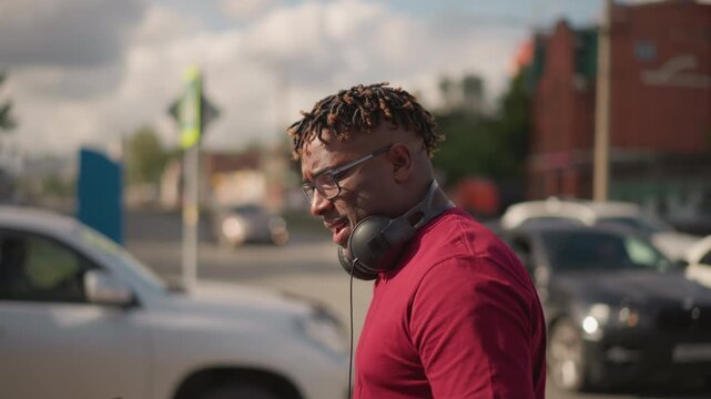 Black DJ cooling down after set, side profile by parking lot, headphones around neck, checking laptop, wiping brow, thoughtful expression, urban traffic, soft sunlight, behind scenes producer mood,