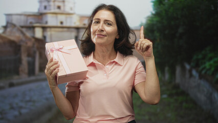 Fototapeta premium Woman holding pink gift box points finger upward on cobblestone street near historic building; celebration gratitude joy.
