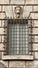 Architectural detail of historic stone window with iron grid and lion sculpture decoration on building facade in Rome, Italy.