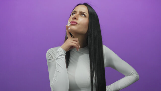 Woman pondering over isolated purple background wearing white shirt showing thoughtful expression.