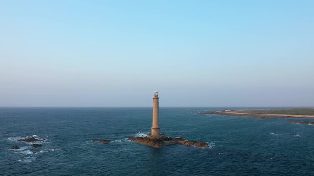 A solitary lighthouse stands on rocky islets surrounded by the sea at Cap de la Hague, Normandy, France, under a clear blue sky. The rugged coastline and open water create a dramatic maritime scene.