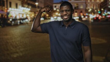 Fototapeta premium Young black man flexes bicep and smiles while wearing glasses on a busy city street at night; confidence strength joy.