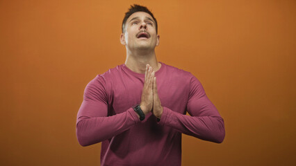 Obraz premium Hispanic man with mustache presses hands together in prayer, head tilted up in a studio with orange backdrop; hope faith gratitude devotion.