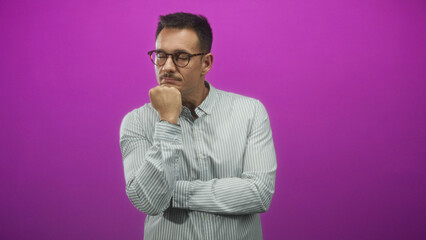 Fototapeta premium Man with mustache and glasses, hand on chin thinking pose in studio with purple backdrop; thoughtful reflection.