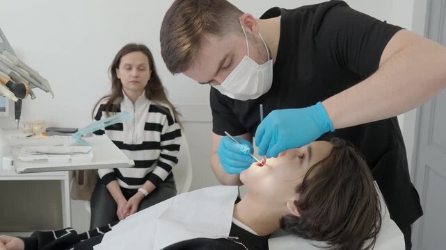 Pediatric dentist examines child oral cavity with probe and mirror. Mother watches in background while routine checkup focuses on caries prevention and painless medical treatment.