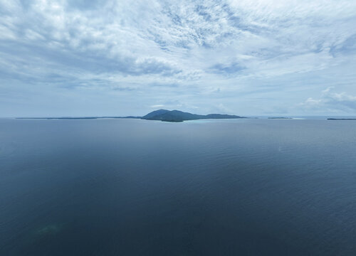 Aerial view of a distant, verdant island rising from the tranquil, deep blue sea under a soft, cloudy sky, Karimunjawa, Central Java, Indonesia.
