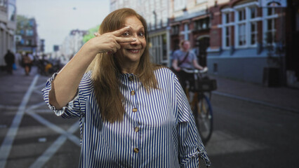Fototapeta premium Woman senior hispanic shows two fingers over eye with hand visible on urban street, striped blouse and slight smile; playful confidence.