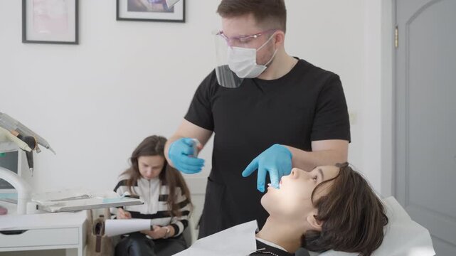 Pediatric dental surgeon removes child primary tooth with surgical forceps. Painless oral surgery procedure involves local anesthesia and biting gauze swab for bleeding control.
