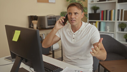 Man on phone pointing at computer screen while seated at desk in an office building; frustration...