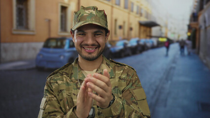 Fototapeta premium Young hispanic man in camouflage uniform energetically claps hands on busy urban street under bright sunlight; pride.