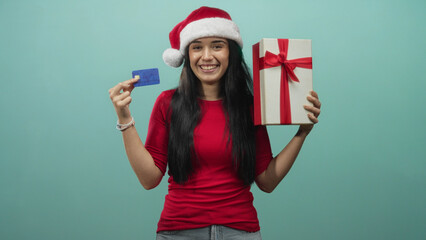 Fototapeta premium Woman holding a creditcard and a large giftbox smiles while wearing a santa hat in a studio; joyful holiday gifting.