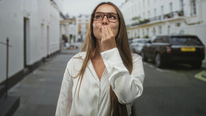 Fototapeta premium Woman with glasses and long hair in a white shirt blowing a kiss with both hands near mouth on a narrow city street lined with parked cars; playful joy.