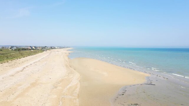 Wide sandy shoreline and calm blue sea under clear sky at Utah Beach, Normandy. Coastal houses line the edge of the tranquil beach landscape.