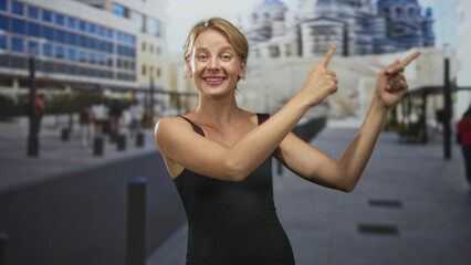 Fototapeta premium Woman pointing both index fingers with bare arms visible in black sleeveless dress on a city street near a modern building and cathedral; joy invitation.