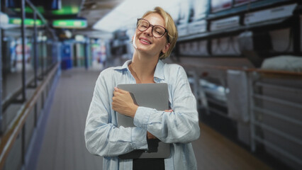 Fototapeta premium Woman hugging tablet to chest, arms crossed and smiling in airport corridor; contentment comfort ease.