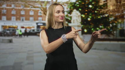 Fototapeta premium Woman points finger to the right on a city street beside a decorated christmas tree and stone statue; cheerful holiday spirit.