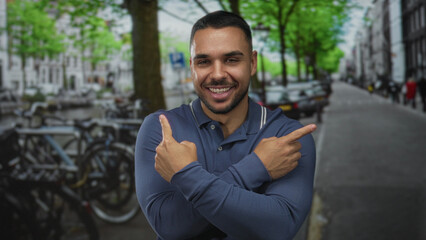 Fototapeta premium Smiling hispanic man pointing index fingers across his chest on city street near parked bicycles and cars; choice confidence.
