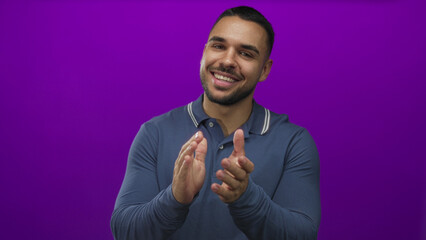 Fototapeta premium Young hispanic man wearing a blue polo shirt claps hands with a broad smile in a purple studio setting; celebration.