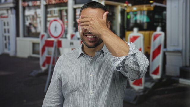 Young man in striped shirt covering face with hand on busy city street near red barriers; embarrassment.