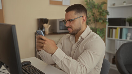 Fototapeta premium Man squints at smartphone screen while gripping device at desk with computer monitor in building; frustration.