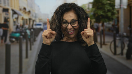 Fototapeta premium Middle aged hispanic woman points both index fingers upward on street while smiling amid bustling storefronts; hope optimism aspiration joy.