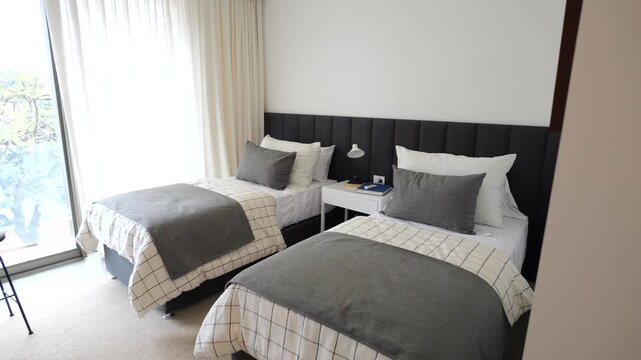 Two twin beds with gray sheets and pillows. The room is very clean and organized. The bed is next to a window, which lets in natural light