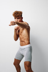 Naklejka premium Athletic young man with curly hair in gray shorts practicing boxing stance and punches for fitness training and workout on white background in studio. Sport, exercise and strength.