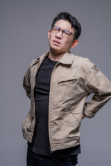 Obraz premium Asian man suffering from lower back pain holding lumbar area, spine injury and muscle strain concept, healthcare problem, tired male with glasses feeling discomfort, studio portrait isolated backgroun