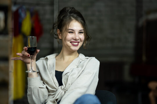 Young caucasian beautiful brunette woman sitting by the table at home or restaurant holding a glass of red wine talking and gesturing with hand. smiling happy girl wearing shirt