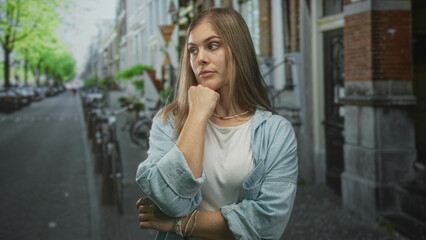 Fototapeta premium Woman with hand on chin standing in a leafy city street lined with bicycles and brick houses; contemplation.