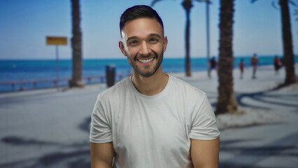 Fototapeta premium Hispanic man smiling at the seaside promenade with palm trees lining the beach as people leisurely walk along the outdoor path enjoying the sunny day by the sea
