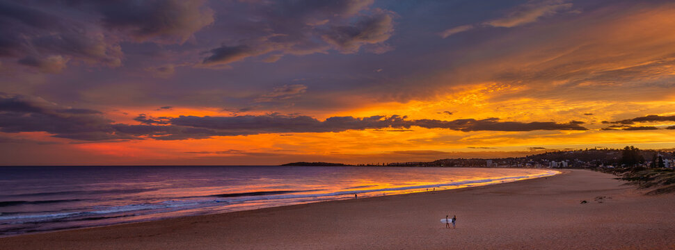 Wide panoramic view of Narrabeen Beach at sunset with surfers on sand. Dramatic orange sky over Northern Beaches coastline in Sydney Australia. Perfect for travel and vacation marketing.