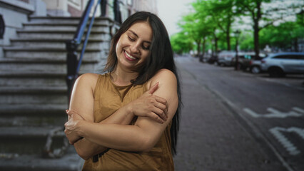 Fototapeta premium Woman hugs herself with bare arms on a city street by stone stairs, smiling in a casual pose and closing eyes; self care joy.