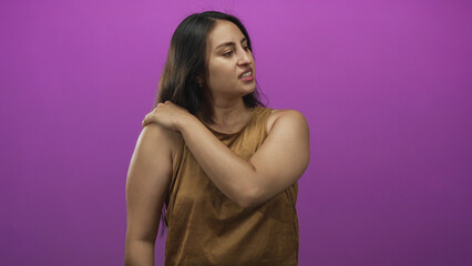Fototapeta premium Woman touching her shoulder and wincing in a purple studio setting with exposed upper arm and hand on shoulder; pain.