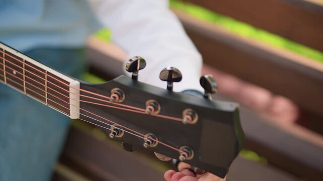 closeup acoustic guitar tuning on bench, young guitarist adjusting tuning pegs with ringed fingers, denim jeans visible, sunlight dappling wood, relaxed introspective practice session focused