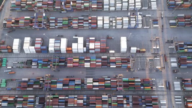 Aerial drone footage showing general container terminal activity at a busy industrial cargo port during evening hours. Stacked shipping containers, cranes and freight logistics infrastructure 