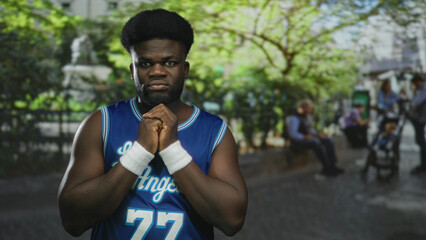 Man with hands clasped at chest wearing a blue basketball jersey and wristbands on a street near...