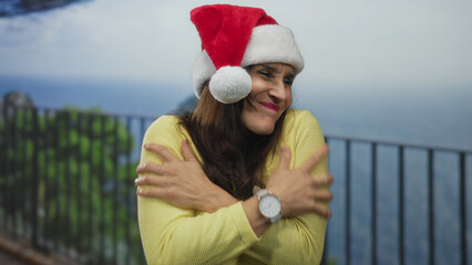Fototapeta premium Hispanic woman wearing santa hat and yellow sweater hugging her arms on seaside promenade; chill winter holiday cheer.