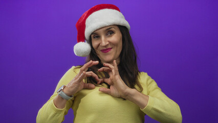 Fototapeta premium Woman in santa hat smiling over isolated purple background making heart gesture representing holiday spirit and festive joy with yellow sweater.