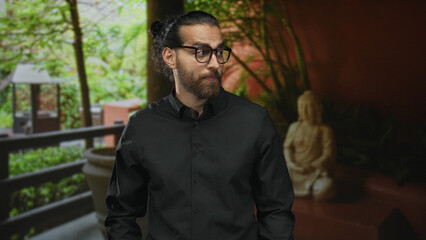 Obraz premium Man with glasses and beard standing in a hotel building lobby, wearing black shirt and gazing sideways near balcony and statue; contemplation.