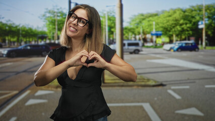 Fototapeta premium Woman with glasses and black top forming a heart with both hands and pointing a finger toward viewer at a tree lined urban street intersection with cars and crosswalk; affection joy.