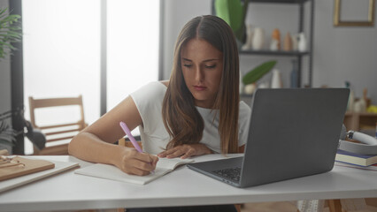 Woman writing in notebook with laptop on desk in studio, leaning forward, pen in hand, focused...