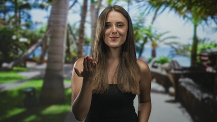 Young hispanic woman with long brunette hair extends palm, beckons with open hand wearing black strapless top in studio; welcoming warmth.