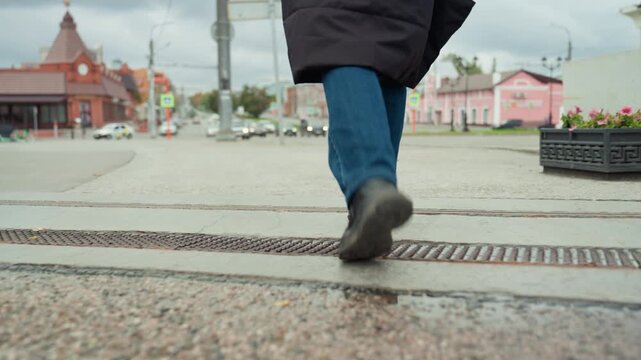 closeup walking feet across plaza urban morning commute vibe brisk steps pavement detail colorful buildings in background coat hem swaying jeans black shoes movement rhythm loopable stock clip