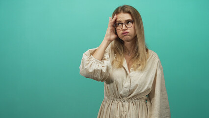 Obraz premium Blonde woman with hand to temple and glasses, beige dress against teal studio backdrop showing headache and tired gesture; fatigue headache.